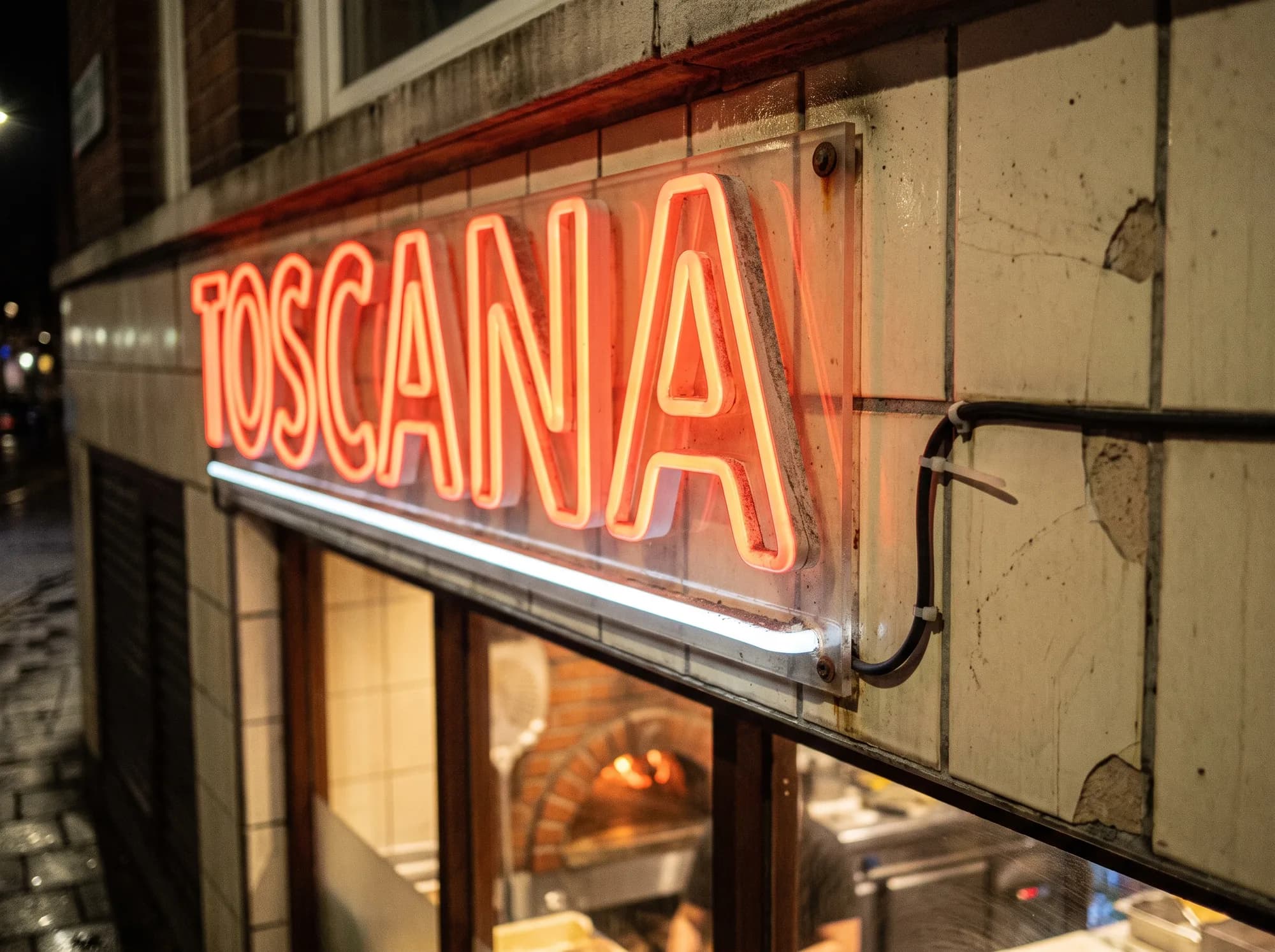 Roter Neon-Schriftzug mit weißer Unterstreichung auf der gefliesten Blende einer Pizzeria bei Nacht